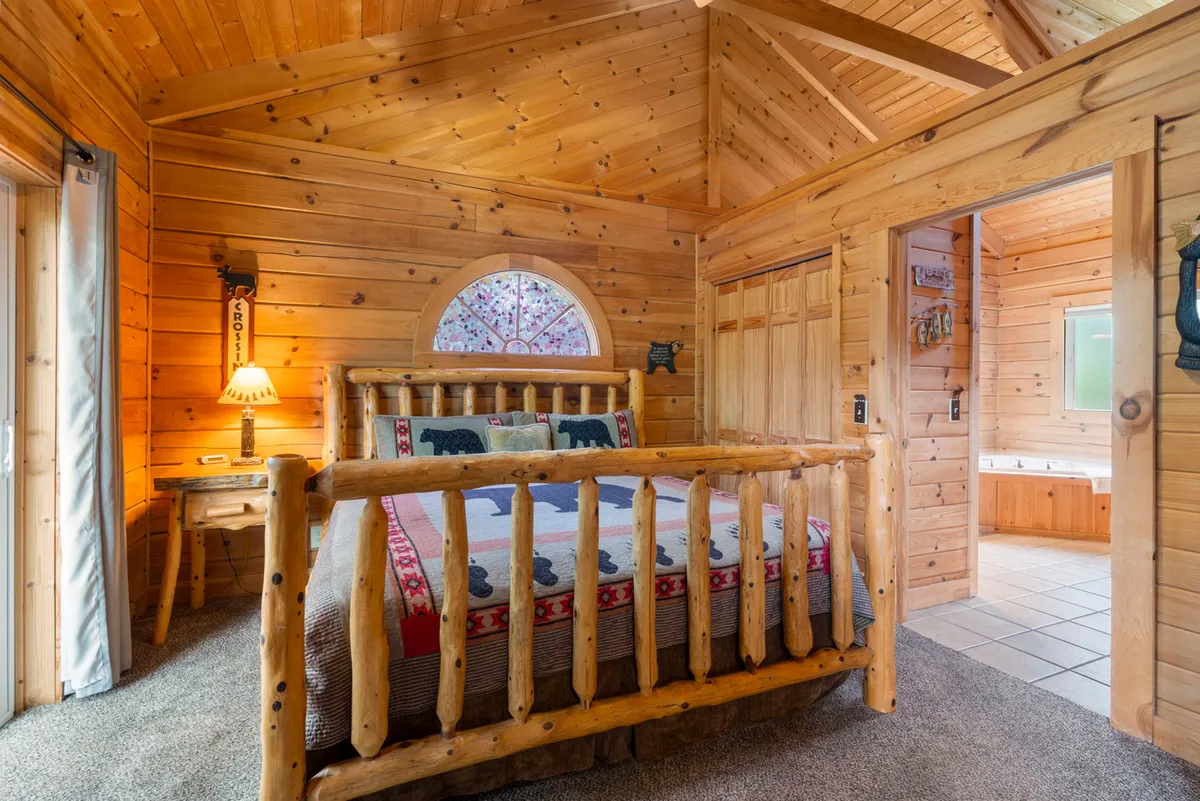 A cozy master bedroom featuring a rustic log bed covered with a decorative quilt, a wooden nightstand with a lamp, and an arched window allowing sunlight to fill the room. Warm wood tones are present throughout, creating a welcoming and restful atmosphere.