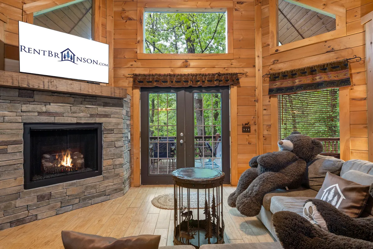 The image showcases a cozy living room with soaring vaulted ceilings, a crackling stone fireplace as the centerpiece, a charming oversized bear plush toy adding a playful touch, double glass doors that open to serene woodland views, and a modern TV for entertainment. The warm lighting and rustic wood accents create an inviting, homey ambiance.