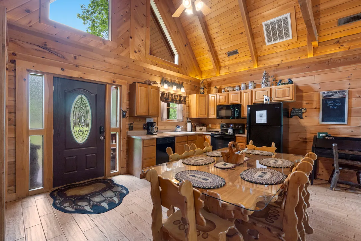 This image captures the inviting interior with a view of the kitchen cabinetry, a central dining table, and rustic ceiling beams. Natural light pours in, highlighting the warm, wood-accented decor and the sense of communal gathering space. The proximity of kitchen and dining areas emphasizes the flow for meal preparation and shared dining experiences.