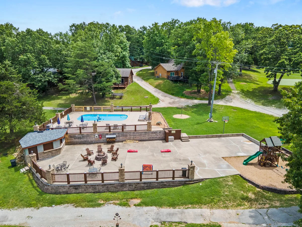 This vibrant exterior scene features a sparkling swimming pool surrounded by lounge chairs and an inviting outdoor seating area. In the background, a cheerful children’s playground and a basketball hoop are visible, all enclosed by lush greenery. The image radiates activity and fun, with spaces for both relaxation and energetic play.