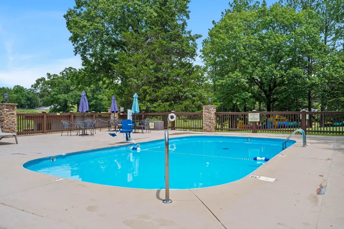 A lively resort swimming pool surrounded by lounge chairs and umbrellas, set amidst tall trees. The atmosphere is cheerful, with guests swimming, sunbathing, and socializing under the open sky.