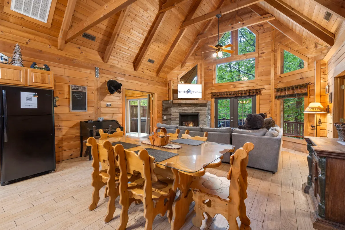 A welcoming open-concept living room with a wooden dining table set for family meals, a cozy stone fireplace as a central feature, a large comfortable sofa, and expansive windows offering serene woodland views. The soft lighting and warm wood accents create an atmosphere perfect for both lively gatherings and quiet relaxation.