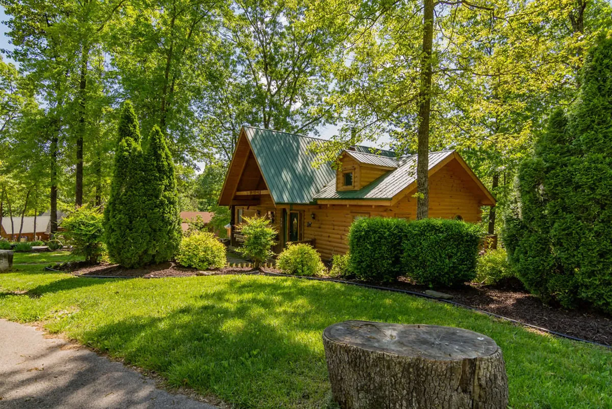 A charming log cabin is set amidst a vibrant green lawn, with tall trees and flowering shrubs offering a serene, natural backdrop. The spacious lawn provides ample room for leisurely strolls or dog walks, and the porch suggests a welcoming spot to relax after outdoor adventures.