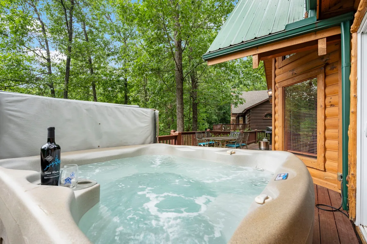 This exterior image features a private hot tub set on a wooden deck, with a bottle of wine nearby. The deck overlooks tranquil wooded hills, bathed in sunset light, creating an intimate and serene atmosphere perfect for couples. The scene suggests relaxation, nature immersion, and special shared moments.