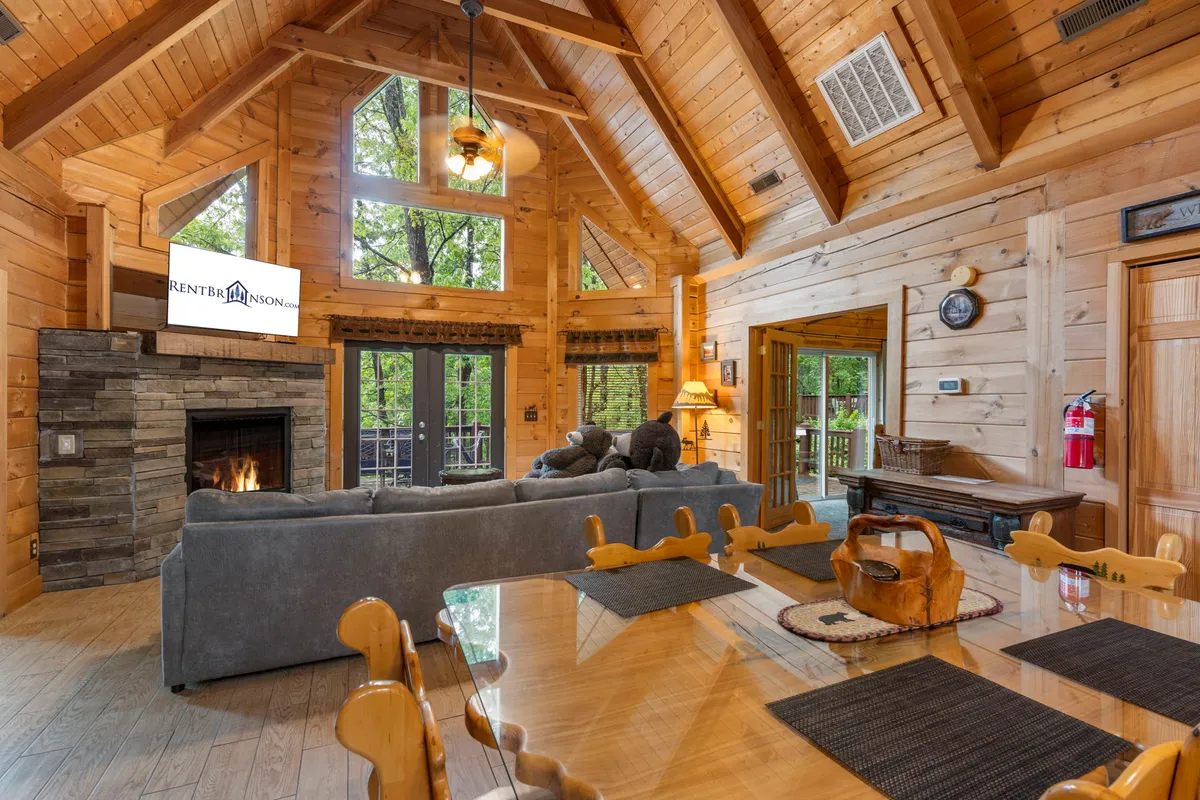 A spacious living and dining area featuring a substantial stone fireplace, a large window filling the space with natural light, and a dining table set for gatherings. The overall feel is warm, welcoming, and perfect for group activities like game nights or shared meals.