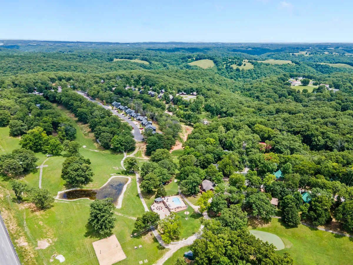 A panoramic outdoor view with a lush green landscape, swimming pool, pond, walking paths, and log cabins. The scene is peaceful, highlighting multiple resort features and abundant opportunities for hiking and nature walks.