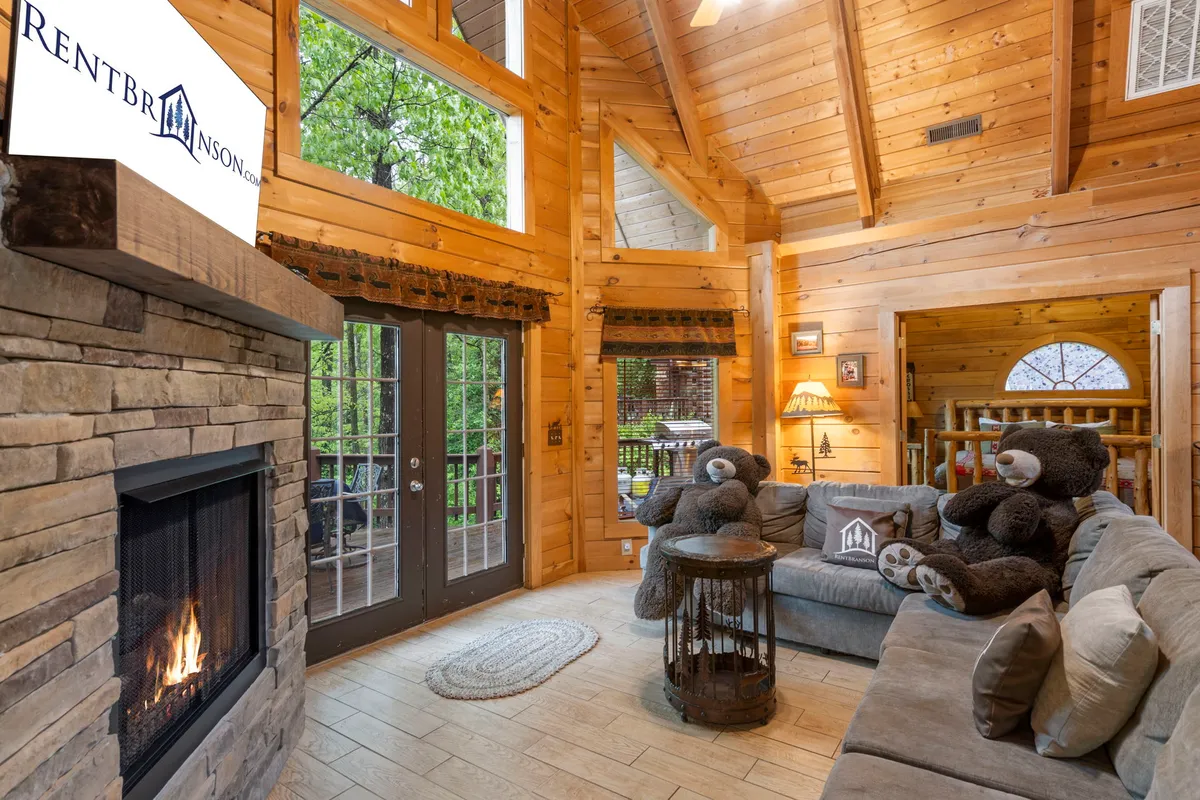 A comfortable living space centered on a stone fireplace, flanked by large windows that let natural light pour in. The sofa is adorned with bear-themed decorations, and the room exudes a warm and inviting atmosphere ideal for both family gatherings and quiet moments.